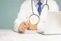 Doctor in a white coat holding a magnifying glass in front of a laptop computer.
