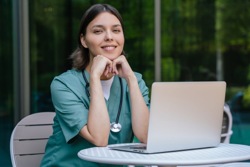 Caregiver in scrubs sitting at a small table and using a laptop, appearing calm and focused in an outdoor work setting.