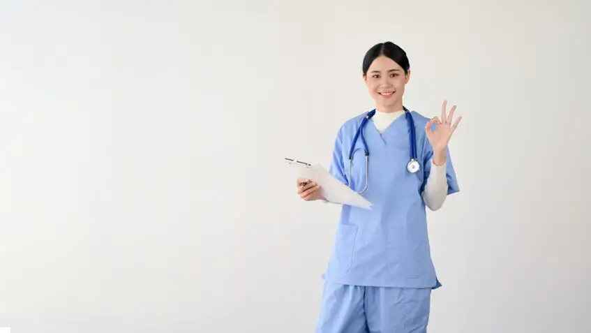 Healthcare professional in blue scrubs holding a clipboard and making an “OK” gesture, standing against a plain white background.