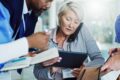 A healthcare team collaborates around a tablet and notes, with a senior professional multitasking on the phone while reviewing patient or operational information.