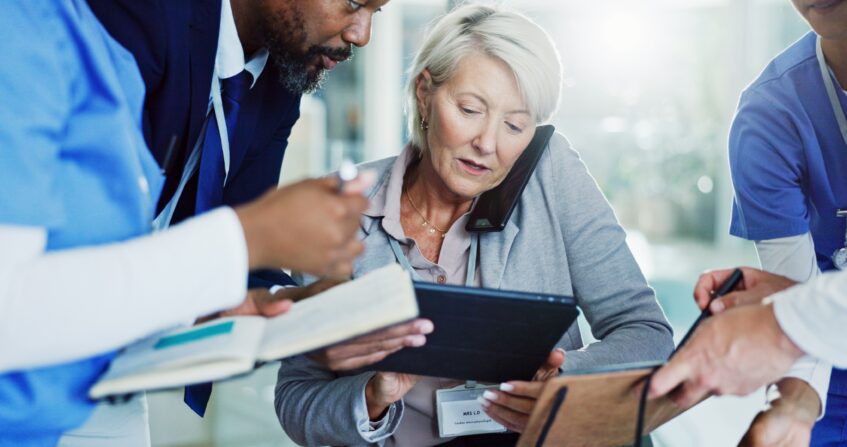 A healthcare team collaborates around a tablet and notes, with a senior professional multitasking on the phone while reviewing patient or operational information.