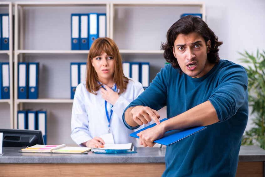 A frustrated man gestures urgently at a clipboard while a concerned administrative staff member stands behind a reception desk, illustrating tension caused by operational strain and administrative breakdown.
